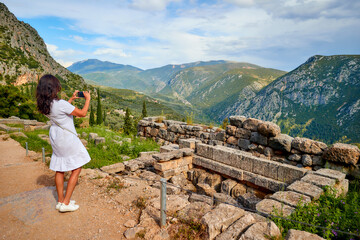  A long-haired woman, wearing white dress  taking pictures of the Apollo Temple ruins, illuminated by sunset. Delphi, Greece.