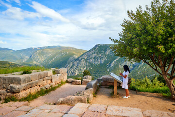 A long-haired woman in a white dress photographing the ancient temple complex of Athena Pronaia in Delphi. Sunny day, blue sky. Archaeological site, UNESCO World Heritage Site, Delphi, Greece.