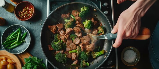 A person is shown from a top view stirring broccoli and meat in a skillet on a stovetop. The...
