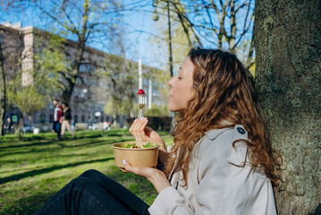 Fototapeta premium A young woman savors a bite of strawberry atop her salad, sitting serenely against a tree in a bustling city park. The moment captures the blend of urban life and the quest for wellness.