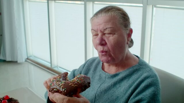 Aged woman pensioner eating chocolate donut and sweet food, unhealthy eating for retired people. Happy grandmother eating food with sugar while her grandchildren out.