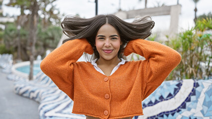 Portrait of a smiling young hispanic woman outdoors, with flowing hair and wearing an orange sweater.