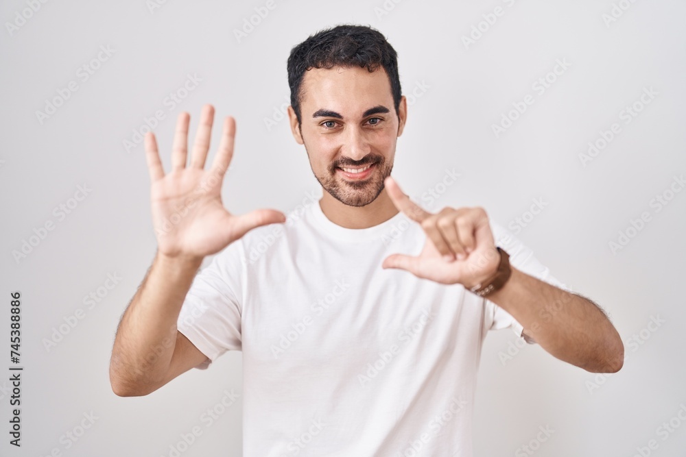 Sticker Handsome hispanic man standing over white background showing and pointing up with fingers number seven while smiling confident and happy.