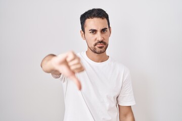 Handsome hispanic man standing over white background looking unhappy and angry showing rejection and negative with thumbs down gesture. bad expression.