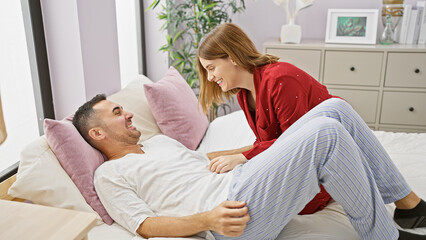 A woman in red leaning over a laughing man in a white shirt on a bed, portraying love in a cozy bedroom setting.