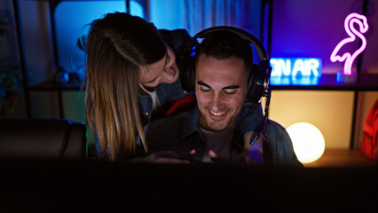 A woman leans affectionately towards a smiling man gaming indoors at night, illuminated by neon lights and monitors.
