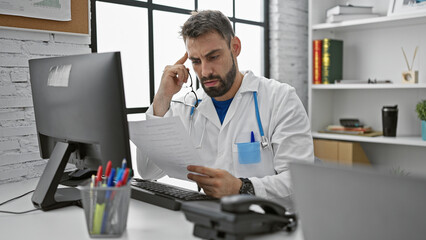 Young and serious hispanic male doctor immersed in his work, reading medical report on computer at...