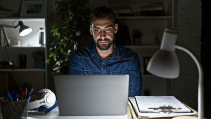 Driven young hispanic man, working long nights at office; focused, serious yet relaxed, this bearded professional navigates business on his laptop.