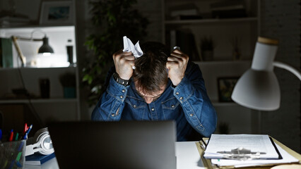 Young handsome hispanic man, seriously stressed out at work, breaking papers over laptop in dim...