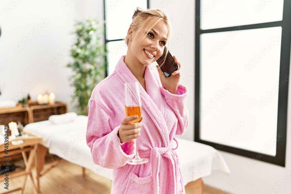 Poster Young caucasian woman wearing bathrobe drinking champagne talking on smartphone at beauty salon