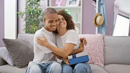 Obraz premium Beautiful couple sitting together on the sofa at home, surprising each other with a lovely birthday gift and confidently smiling as they enjoy a hugging moment in living room background.