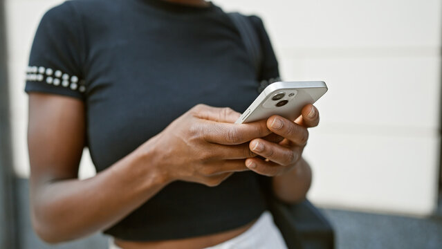Attractive african american woman engrossed in digital conversation, using her phone outdoors, touching the screen to text in an urban city street setting.