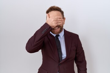 Middle age business man with beard wearing suit and tie smiling and laughing with hand on face covering eyes for surprise. blind concept.