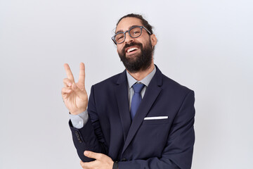 Hispanic man with beard wearing suit and tie smiling with happy face winking at the camera doing victory sign. number two.