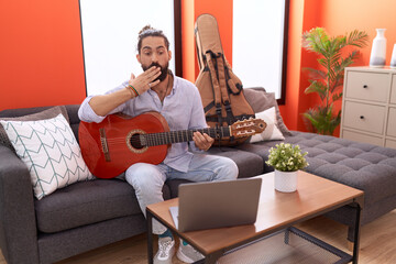 Hispanic man with beard playing classic guitar at home covering mouth with hand, shocked and afraid...