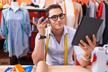 Young hispanic man tailor listening voice message by smartphone using touchpad at atelier