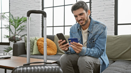 Handsome hispanic man with beard using smartphone and holding credit card in modern apartment...