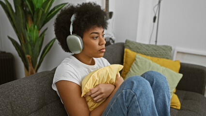 A contemplative african american woman with curly hair wearing headphones indoors on a cozy sofa surrounded by colorful pillows.