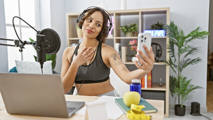 A young woman takes a selfie in a podcast studio setup with laptop, microphone, and headphones.