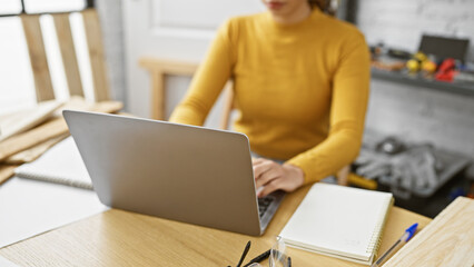 Focused woman working on laptop in a woodworking workshop environment.