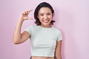 Hispanic young woman standing over pink background smiling and confident gesturing with hand doing small size sign with fingers looking and the camera. measure concept.