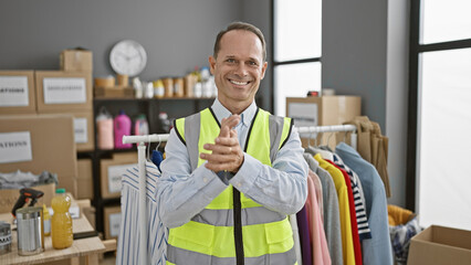 Confident smiling middle age man joyfully clapping hands, volunteering at charity center. handsome adult dons reflective vest, standing amidst donations, oozing unity, altruism in community. - obrazy, fototapety, plakaty