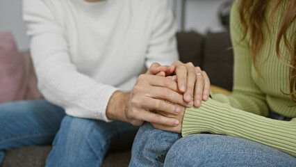 A couple sitting closely on a couch at home, holding hands to show affection and emotional support...