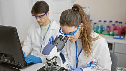 A woman and man, clad in lab coats and safety glasses, analyze results in a brightly-lit scientific laboratory.