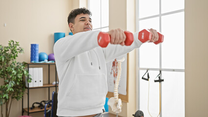 A young adult man with a beard exercises using dumbbells in a bright physiotherapy room, showing a...
