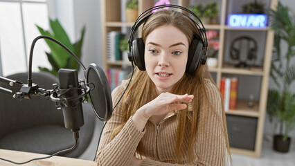 Young blonde woman wearing headphones speaks into a microphone in a modern radio studio.