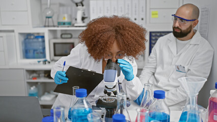 Inside the buzzing lab, two committed scientists, man and woman, analysis partners, seated together, gloved hands busy writing on clipboard, eyes glued to the microscope in serious research - obrazy, fototapety, plakaty