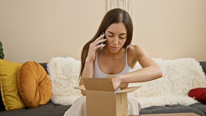 Beautiful young hispanic woman anxiously unpacking delivery parcel while engaged in serious phone conversation at home