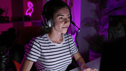 Attractive young hispanic woman, smiling and confident, streaming her gaming adventure, wearing headset in her home gaming room in the dark night, enthralled in a virtual world