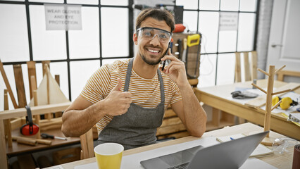 A cheerful young man wearing safety glasses gives a thumbs up while on a call in his well-equipped...
