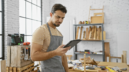 A focused hispanic man with a beard reads a tablet in a bright carpentry workshop filled with tools and wood.
