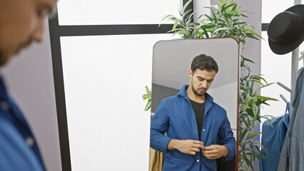 Handsome hispanic man buttoning shirt in a modern dressing room with plants and hats.