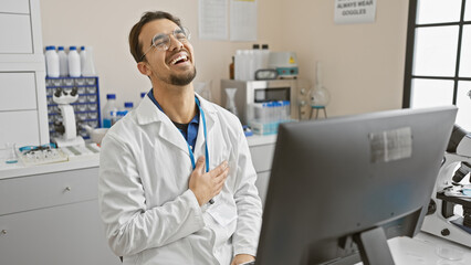 Naklejka na ściany i meble Laughing hispanic man in lab coat sitting at computer in bright laboratory setting.