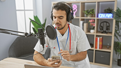 Handsome hispanic man in medical uniform podcasting with microphone and smartphone in radio studio. - obrazy, fototapety, plakaty
