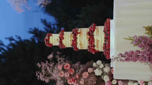 Vertical footage. Close-up beautiful wedding cake decorated with berries on the table with white tablecloth near outdoor wedding arch decorated with white, red, pink flowers at night, slow motion.