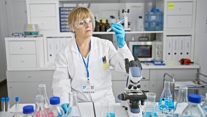 Serious, blonde middle-age woman scientist, taking notes and holding a test tube in a bustling lab,...