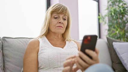 Serious mid-age blonde woman struggles with technology, upset expression as she sits on sofa indoors, typing message on smartphone, engrossed in the problem at hand in her stylish living room