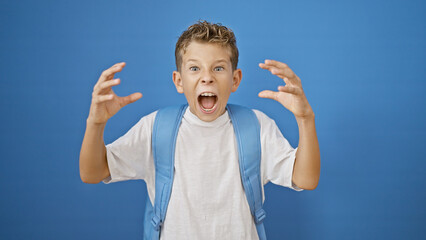 Adorable blond boy student standing, screaming loudly with serious expression over isolated blue...