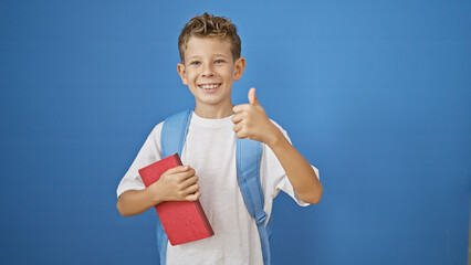 Adorable blond boy student, confidently holding book while giving thumb up ok gesture, smiling widely on isolated blue background, celebrating the joy of learning.