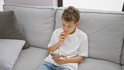 Adorable blond boy, seriously engrossed in watching a video on his smartphone, eating a sweet...