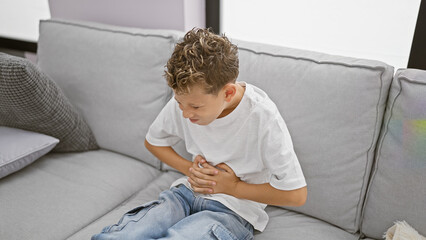 Cute little blond boy suffering from stomach ache, sitting on home sofa with a worried face, touching unsettled stomach