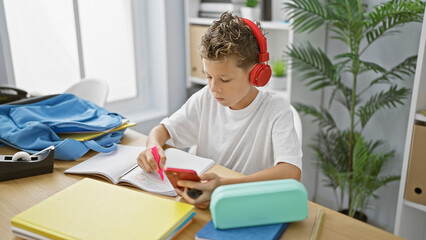 Adorable blond boy student, engrossed in listening to music on his smartphone, diligently drawing...