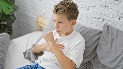 Adorable blond boy, sitting on bed in his pajamas, coughing in the early morning. sick child...