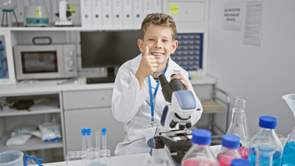 Adorable little blond boy, a budding scientist, giving a thumb up gesture while confidently using a microscope in a medical lab, all smiles amid his exciting scientific discovery.