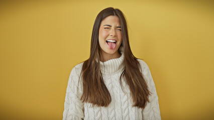 A playful young woman sticks out her tongue, wearing a sweater against a vibrant yellow background.