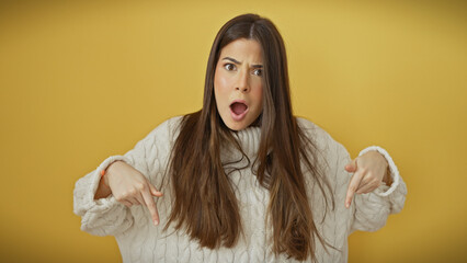 Surprised young hispanic woman with open mouth pointing downwards, wearing a sweater against a...